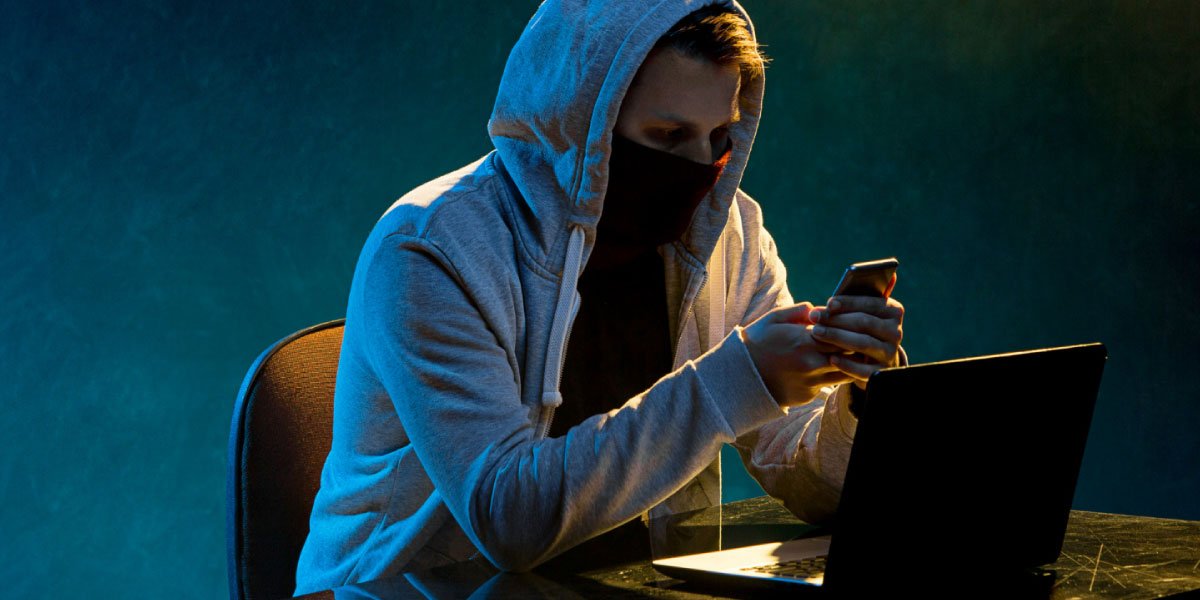 Maskierter Mann, der in einem dunklen Raum mit seinem Laptop vor sich auf sein Telefon schaut