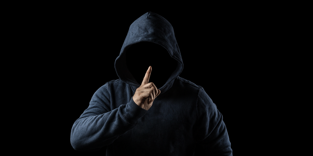 A figure in a dark hoodie with his finger in front of his face with his face in the shadow