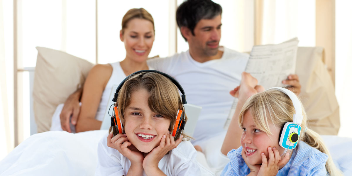Family of four sitting in bed and children having headphones on their heads.