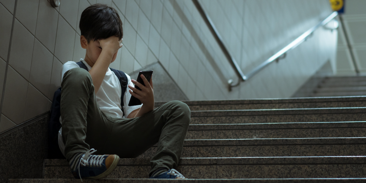 A depressed little boy looking at his smartphone screen sitting at his school staircase