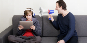 An angry frustrated father yelling into a megaphone at his unconcerned teenage son wearing headphones and using his tablet