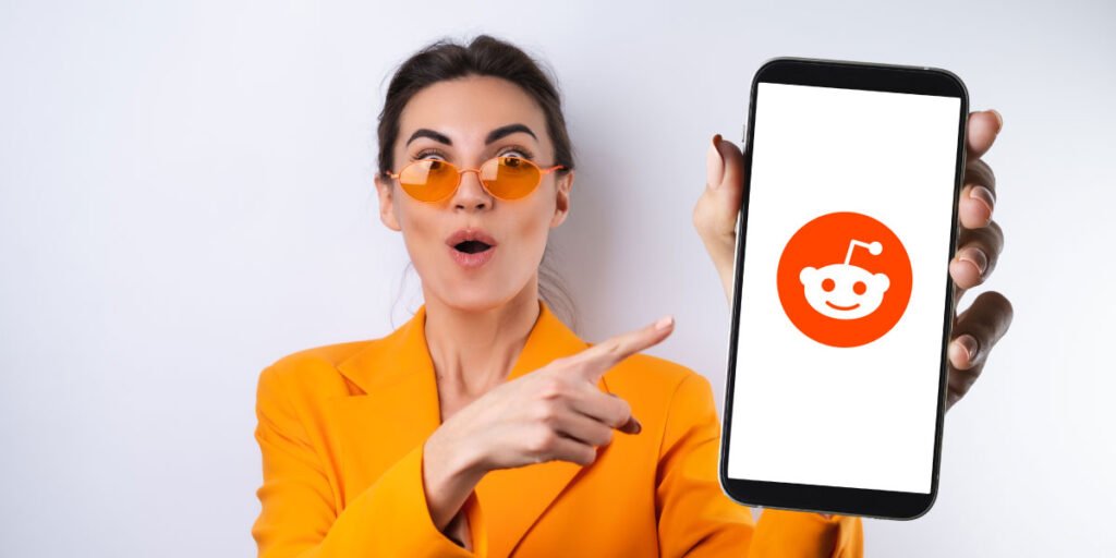 A young woman in an orange outfit, holding her smartphone screen with the Reddit logo up to the camera, wondering if Reddit is safe for her kids