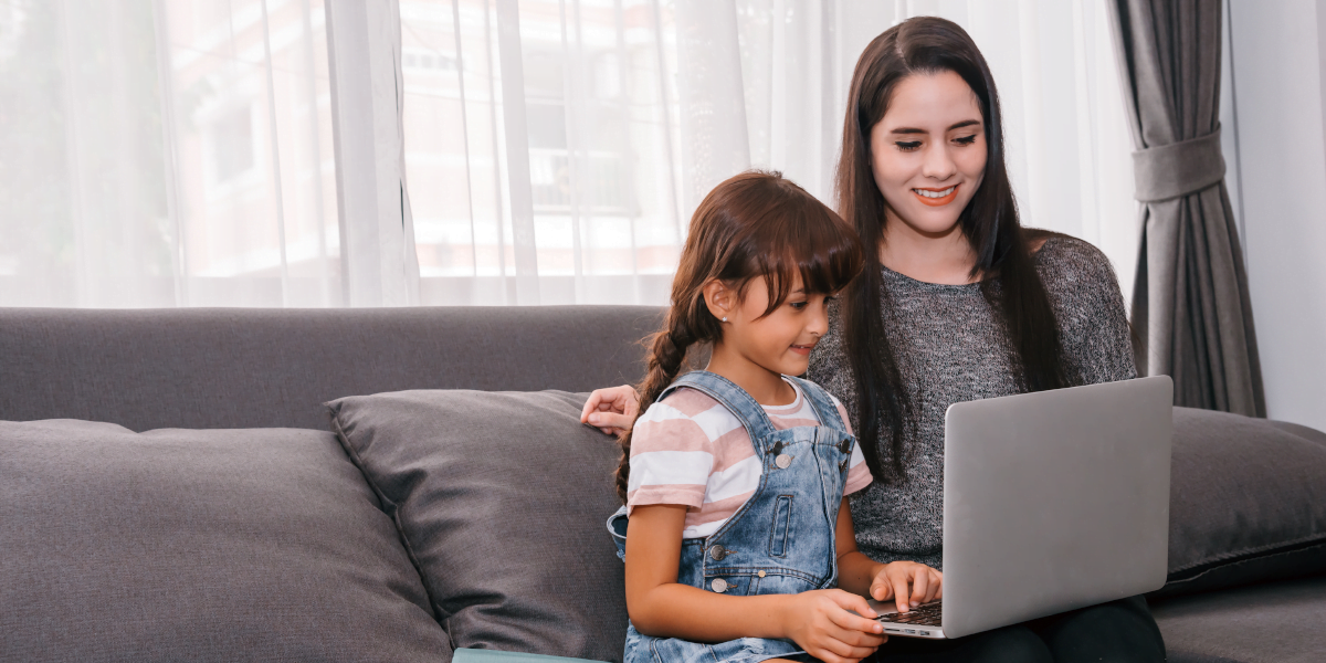 Mother and young daughter sitting on couch with a laptop on their lap while the mother teaches her about online scams