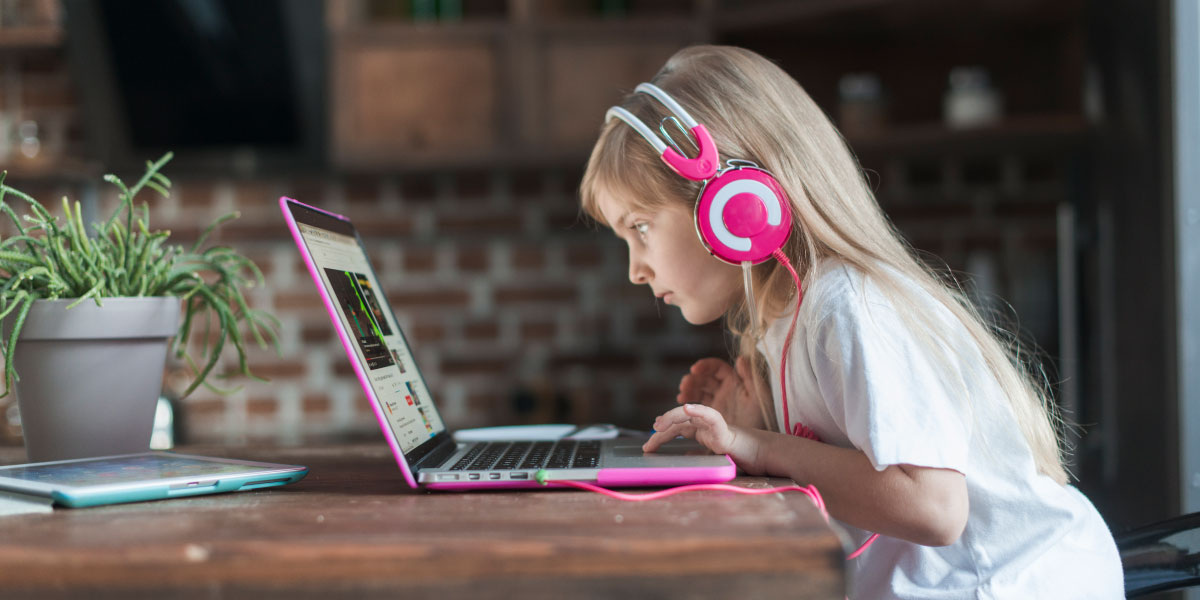 Girl sitting at a table using a laptop and pink headphones.
