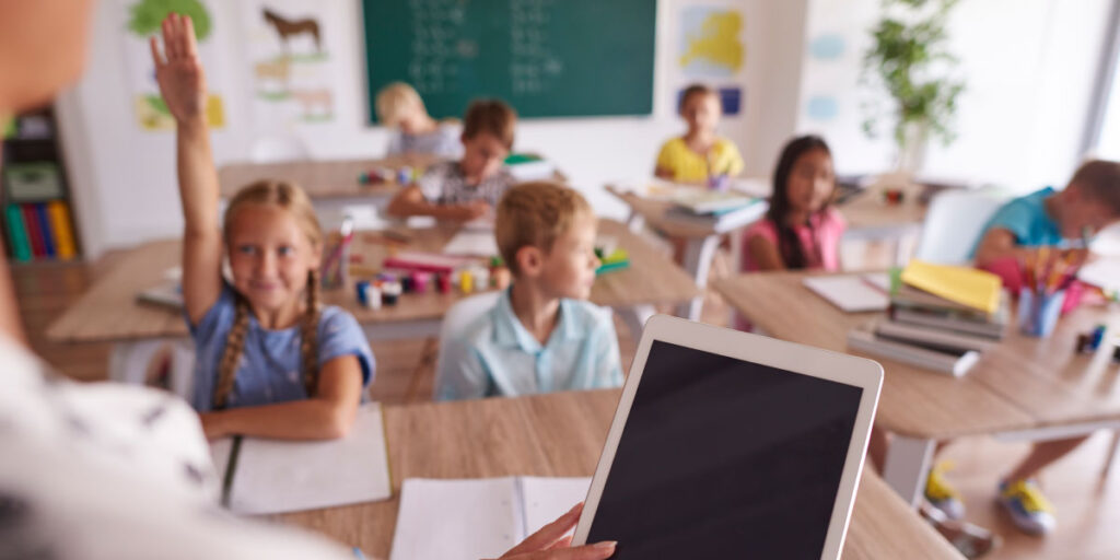 Teacher holding a tablet in a classroom full of kids with one girl holding up her hand