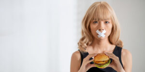 Woman having an X taped on her mouth and having a hamburger in her hand.