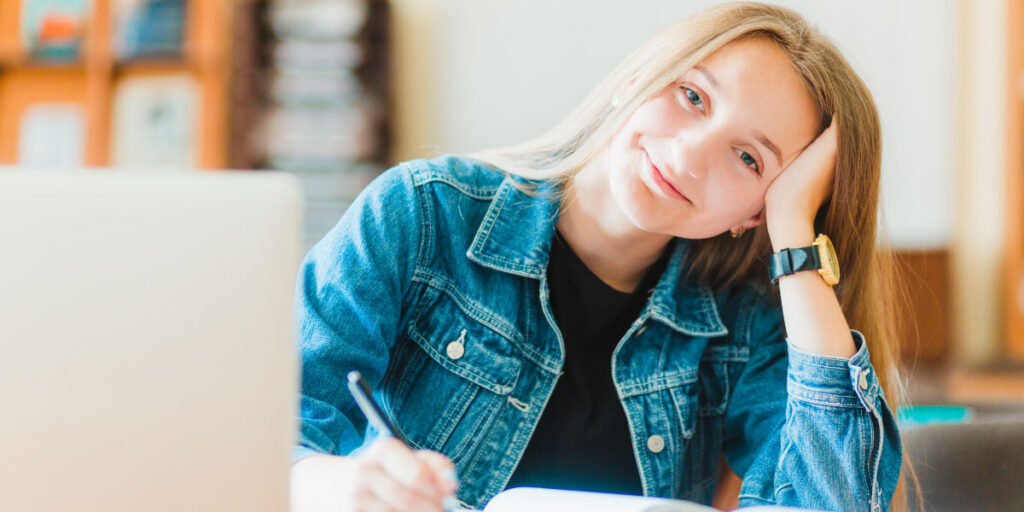 A smiling girl writing in a notebook with a laptop in front of her.