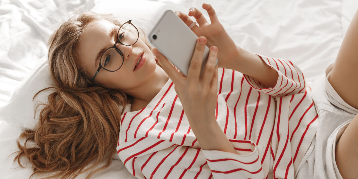Junge lächelnde Frau, die zu Hause auf dem Bett sitzt und mit ihrem Smartphone SMS schreibt