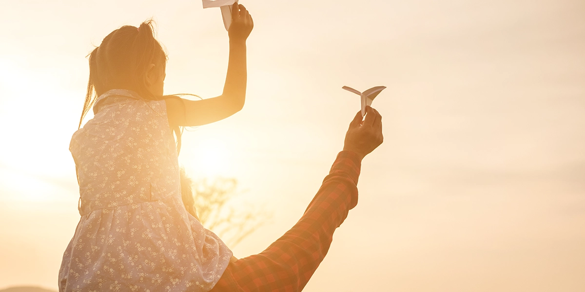 daughter sitting on the neck of her father holding paper airplanes