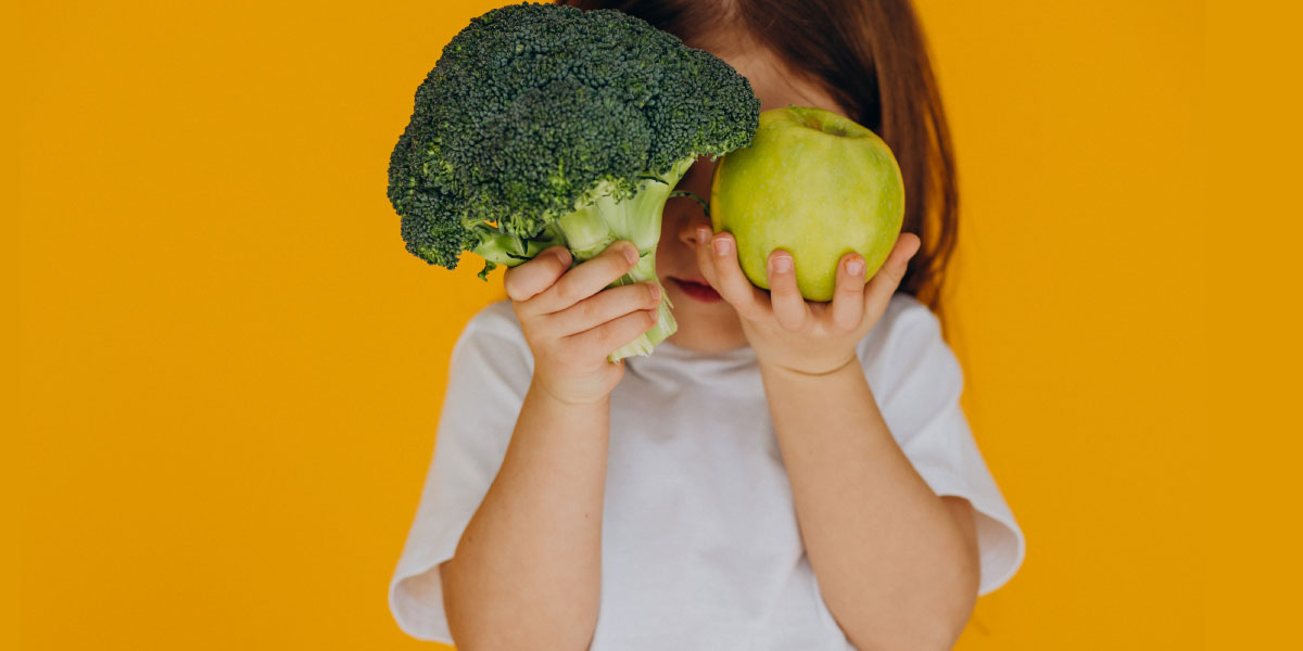 Girl wearing white t-shirt holding broccoli and an apple in front of her face