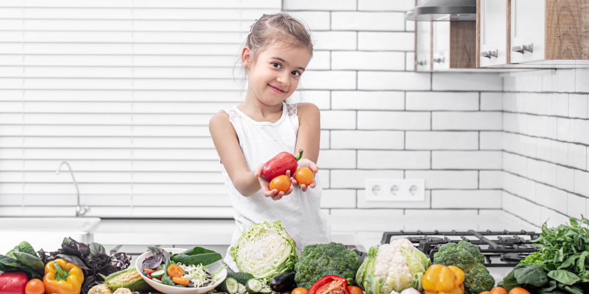 girl standing in front of a counter full of vegetables and holding vegetables in her hand