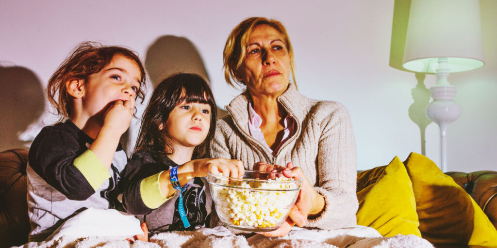Two girls watching a movie with an adult and eating popcorn.