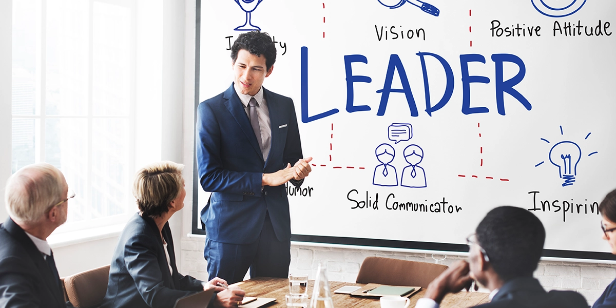 A man talking at a business meeting as if he is a leader. Behind him on a whiteboard, it reads LEADER