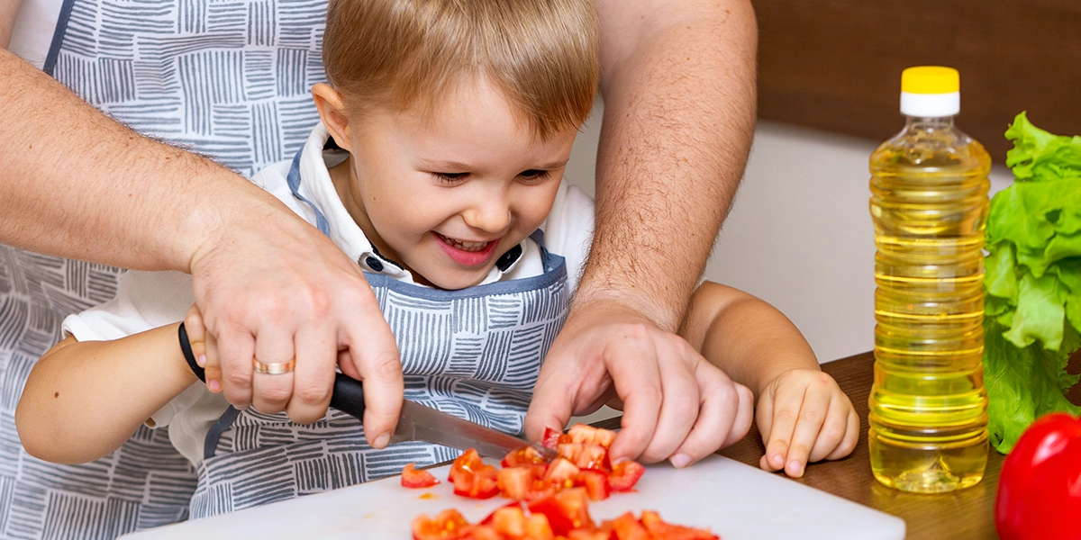 Child being guided to use a knife for chopping tomatoes by an adult