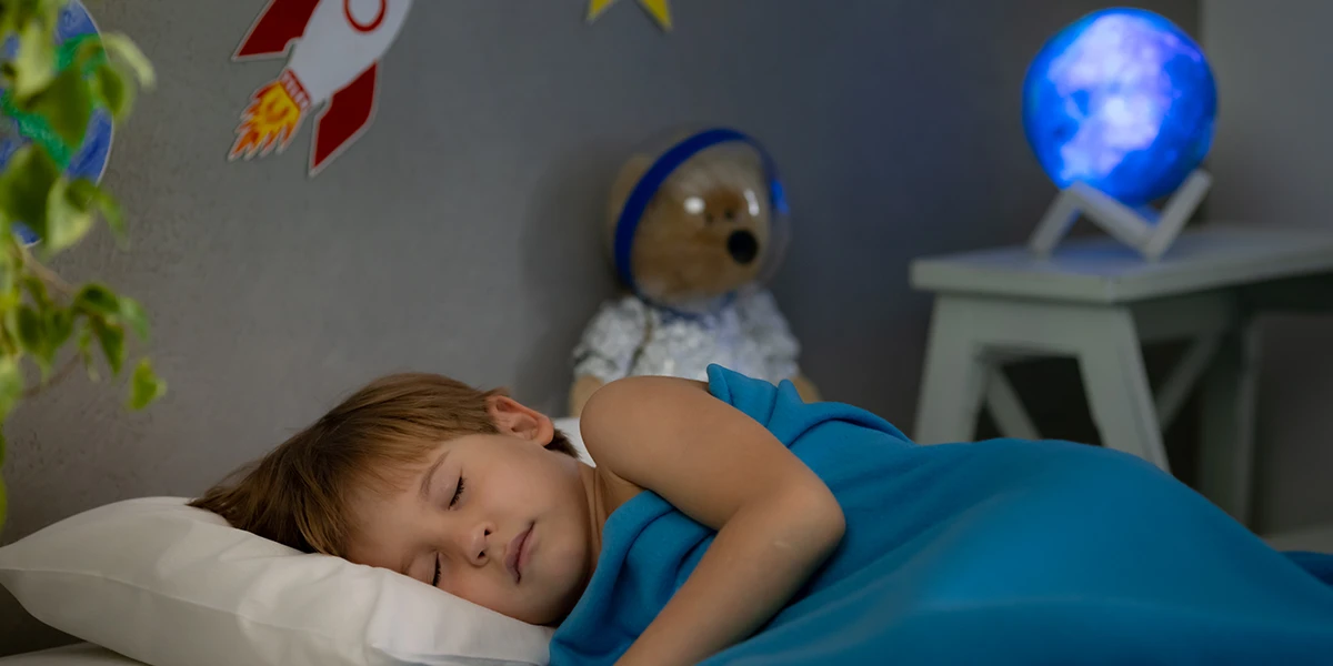 kleiner Junge, der in seinem Bett schläft, mit einem Teddybären und einer Lampe im Hintergrund