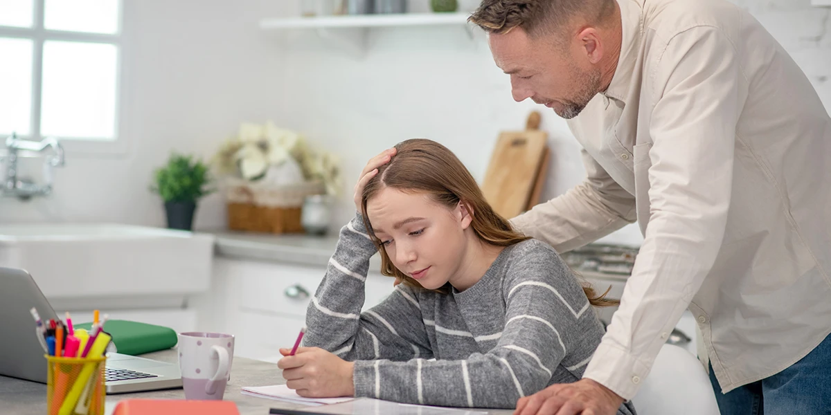 Young girl sitting at table doing homework with father standing behind him
