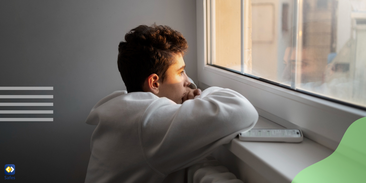 a sad boy looking outside through a window dealing with nature deficit disorder symptoms with his phone next to him