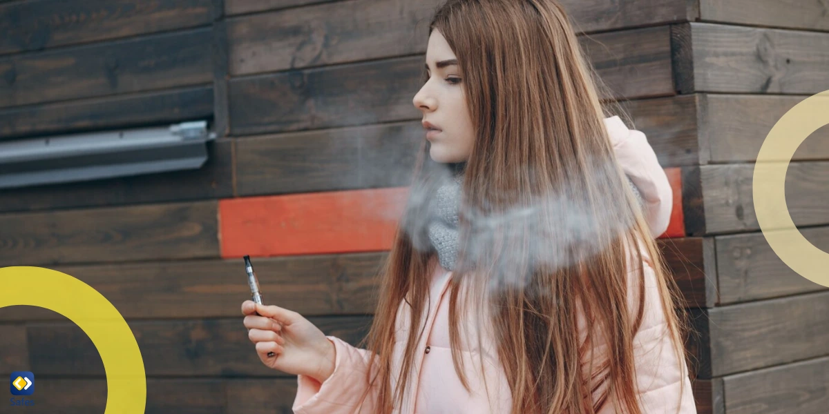 A young woman in a pink jacket and scarf is vaping outdoors, exhaling smoke.