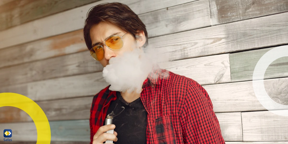 A young man in a red plaid shirt and sunglasses is vaping against a rustic wooden wall, exhaling a large cloud of vapor.